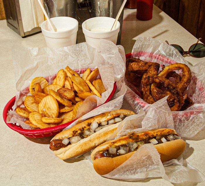Two Detroit Coney dogs with two plates of onion rings and french fries.