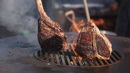 A round metal surface with a round cut out grill in the middle. Glowing fire is seen through the grill grates. Two steak with long bones extending upward are searing on the grill.