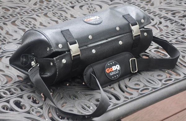 Black leathery cylindrical shaped bag with carrying strap and two shiny buckles to snap it shut.