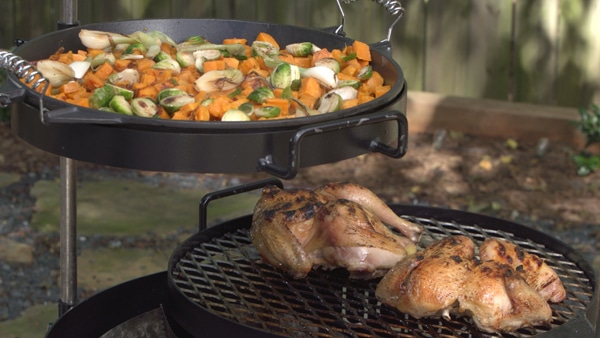 A round cooking grate holding grilled chicken and a round pan filled wih chopped vegetables.