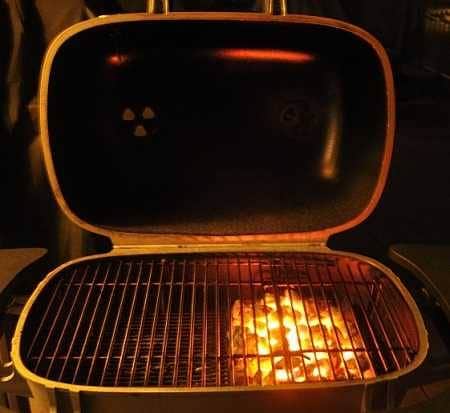 Charcoal grill at night with the lid up The glowing charcoal fire is piled on the right side.