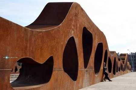 Massive rusted colored, wavy metal outdoor sculpture metal that appears to be about 20 feet high. A man leans against it.